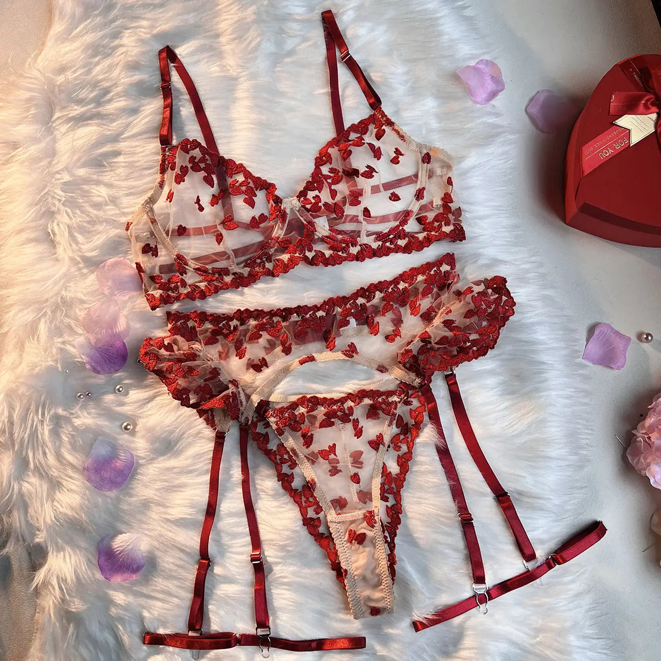 A sheer white lingerie set featuring delicate red heart embroidery, comprising a bralette, thong, and garter belt, artfully displayed on a plush white surface.