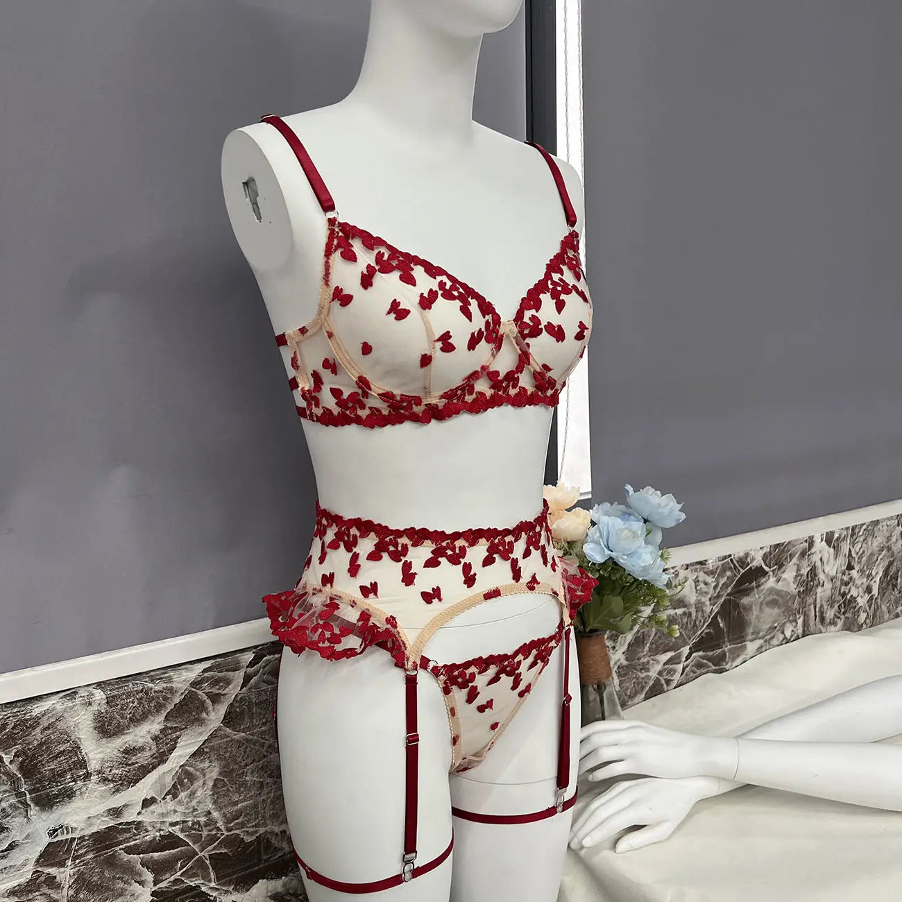 Sheer white lingerie set with red heart embroidery on a mannequin. It includes a bra, garter belt with ruffles, and thong, all featuring red straps and trim.