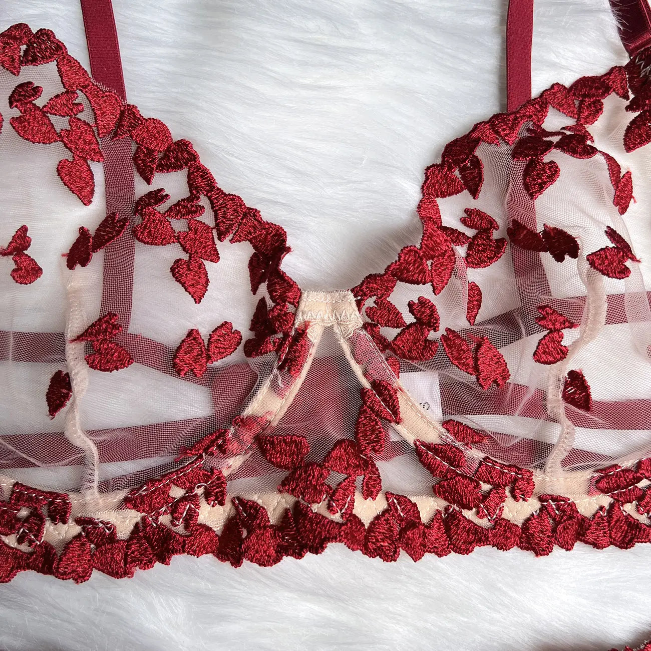 This image features a delicate red embroidered sheer mesh bralette with intricate leaf-like patterns, showcasing its intimate details and luxurious texture against a white furry background.