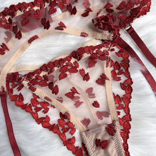 This image features a delicate mesh lingerie piece, likely a garter belt or thong, adorned with vibrant red heart embroidery and subtle beige trim, presented on a soft white furry surface.
