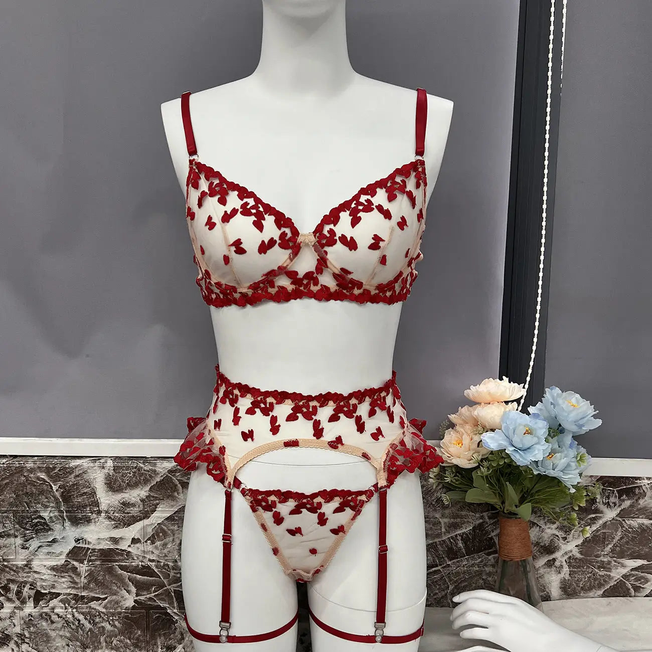 A sheer cream lingerie set featuring red embroidered hearts/butterflies, consisting of a bra, thong, and garter belt with red straps and trim.