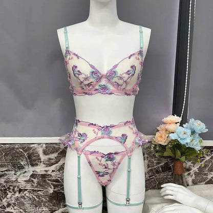 A delicate three-piece lingerie set, featuring a sheer bra, thong, and garter belt. It boasts intricate purple, pink, and teal floral embroidery on a white base, accented with mint green straps.