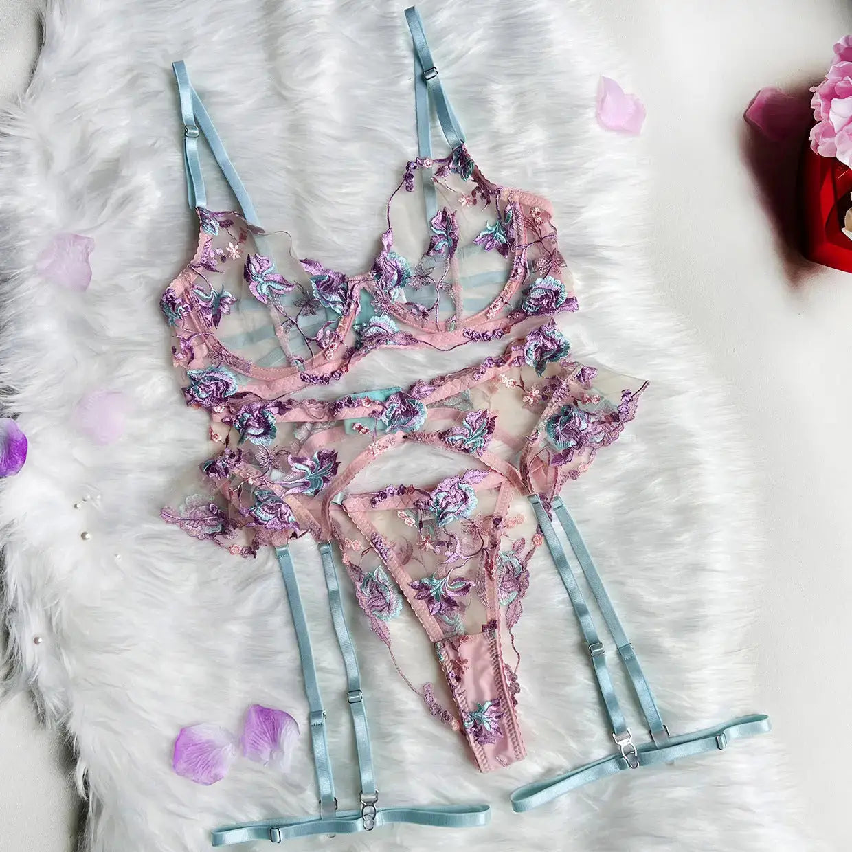 A beautiful pink sheer lingerie set featuring a bra, thong, and garter belt, adorned with intricate purple and blue floral embroidery and light blue adjustable straps, displayed on white fur.