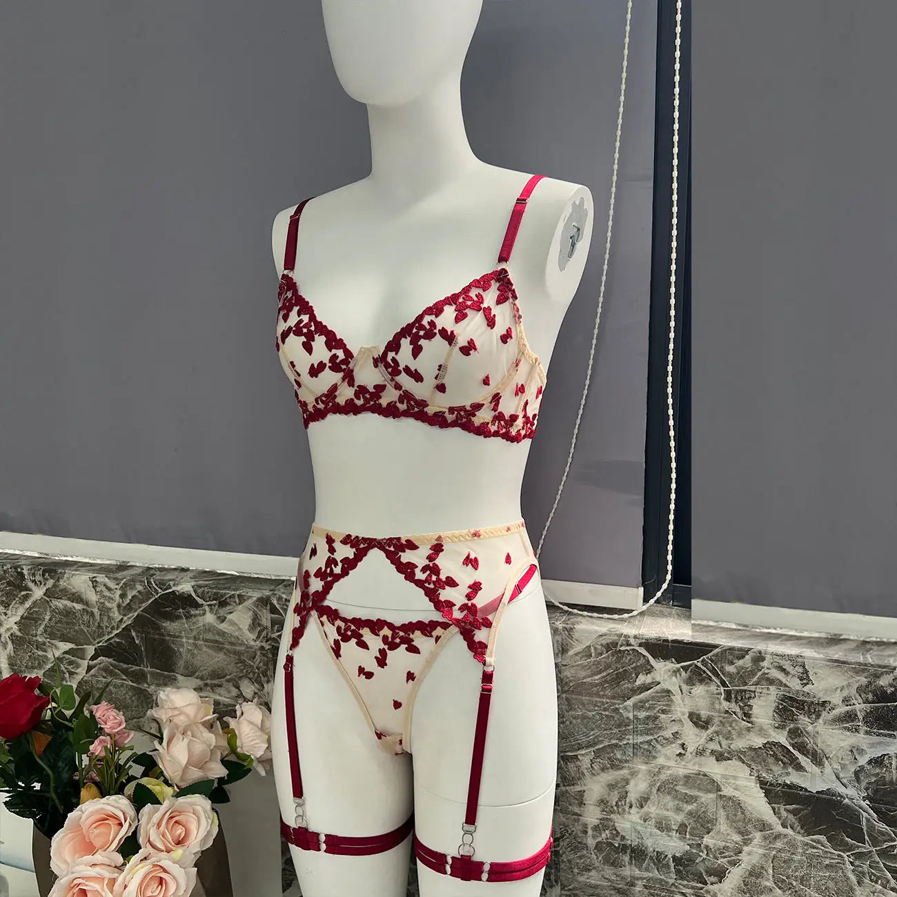 An elegant cream lingerie set on a mannequin, featuring a bra, thong, and garter belt with thigh straps. Adorned with delicate red heart embroidery, perfect for intimate wear.