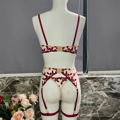 A delicate lingerie set featuring a bra, thong, and garter belt with leg straps. Made of sheer beige fabric with intricate red floral embroidery and matching red straps on a mannequin.