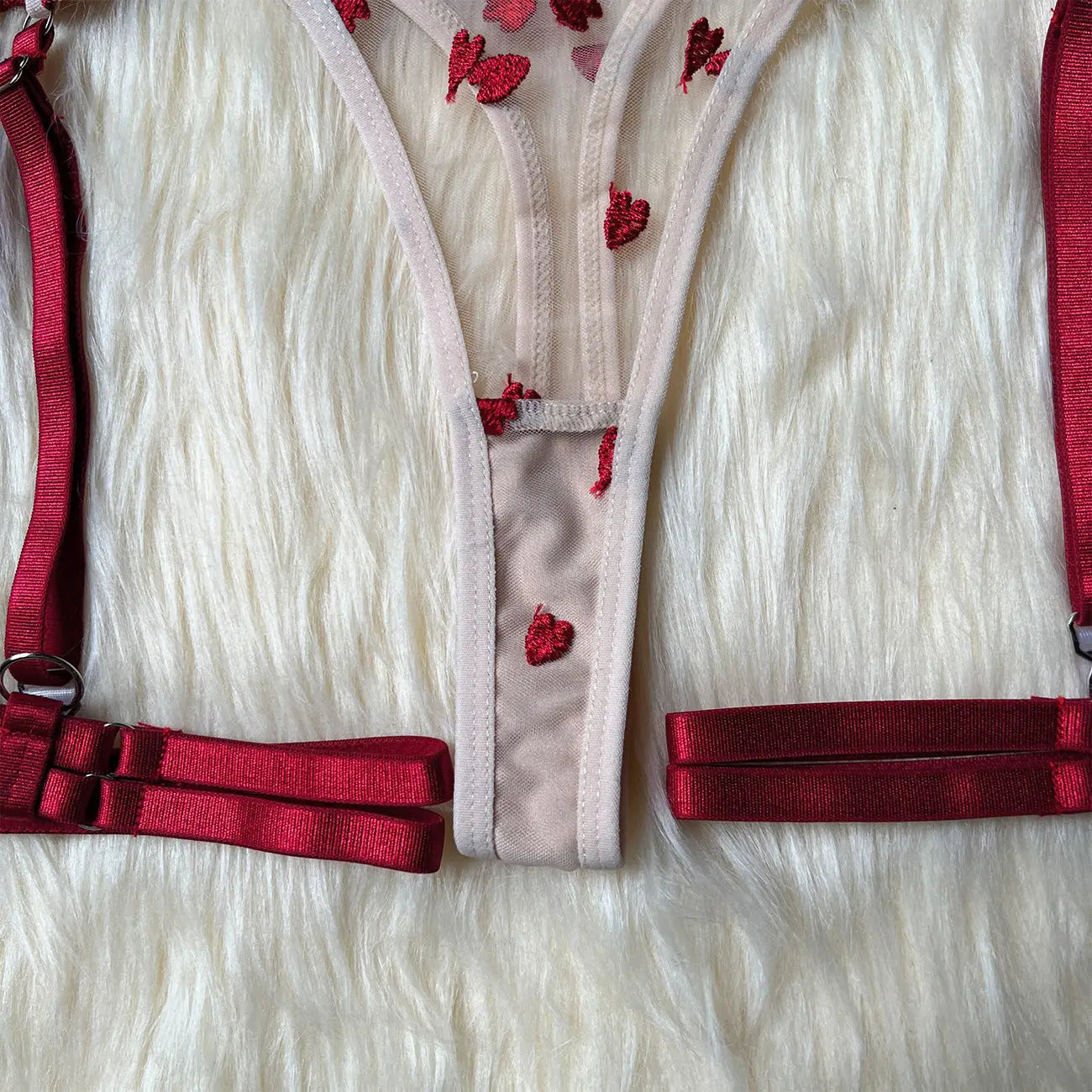 Sheer beige thong featuring embroidered red hearts on delicate mesh, accompanied by matching red elastic straps. This intimate apparel set is displayed on a soft, fluffy white surface.