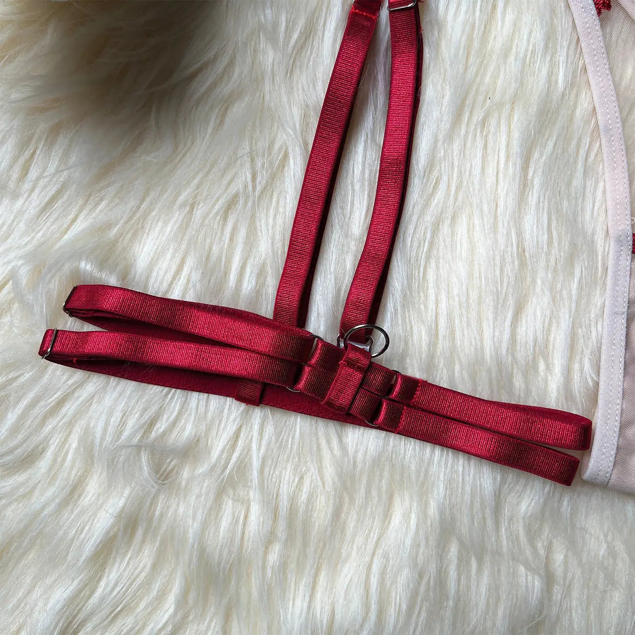 Close-up of a dark red lingerie harness with adjustable elastic straps and a silver O-ring connector, resting on a white faux fur surface. A beige garment with subtle detailing is partially visible.