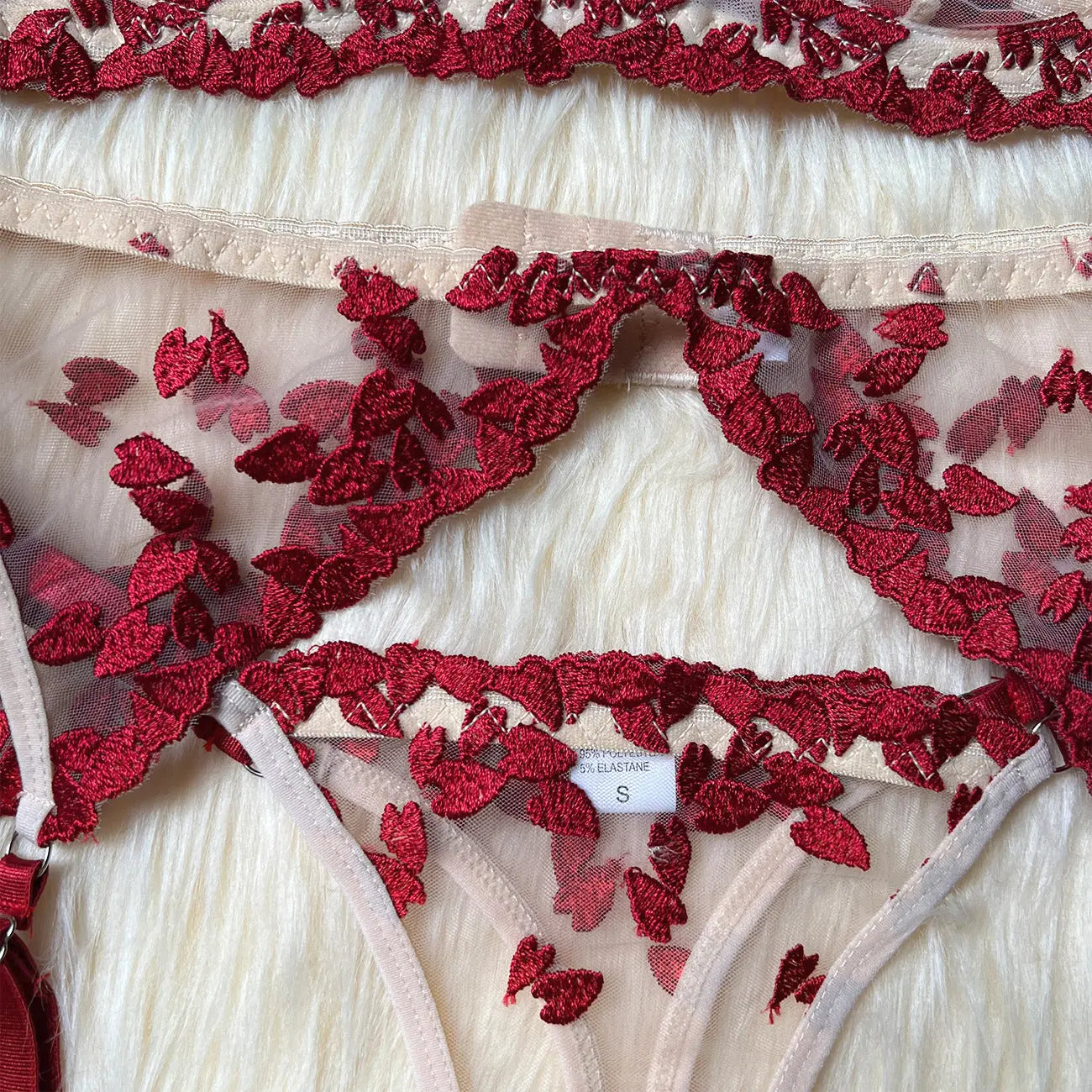 Close-up of sheer beige lingerie featuring delicate red leaf-like embroidery. A visible tag indicates size 'S' and material composition of 95% polyester, 5% elastane, resting on a fuzzy white surface.