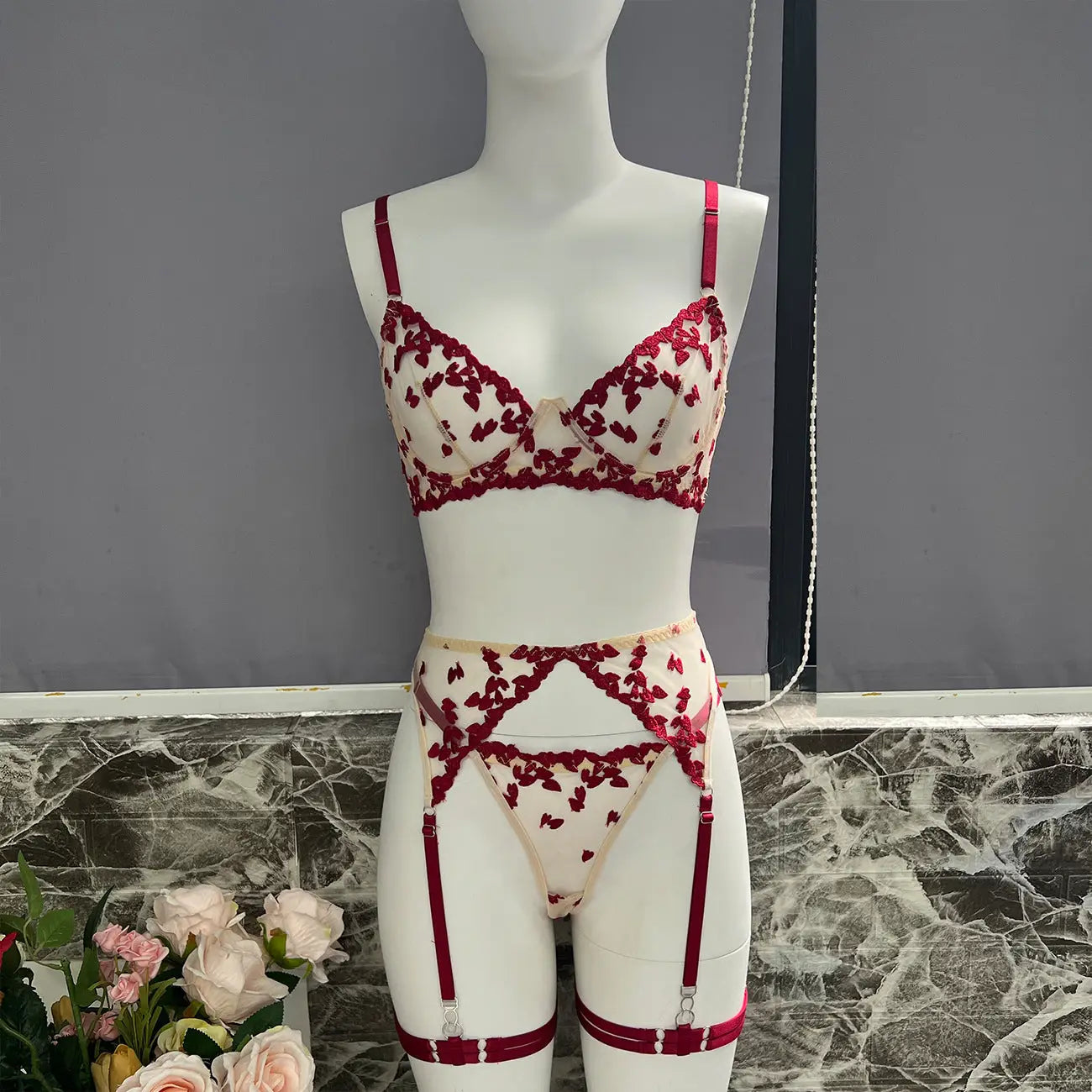 A delicate lingerie set, featuring a bra, thong, and garter belt, adorned with intricate red floral embroidery on sheer, light-colored fabric, displayed on a mannequin.