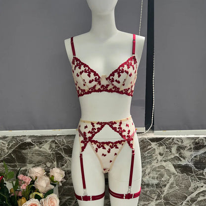 A delicate lingerie set, featuring a bra, thong, and garter belt, adorned with intricate red floral embroidery on sheer, light-colored fabric, displayed on a mannequin.