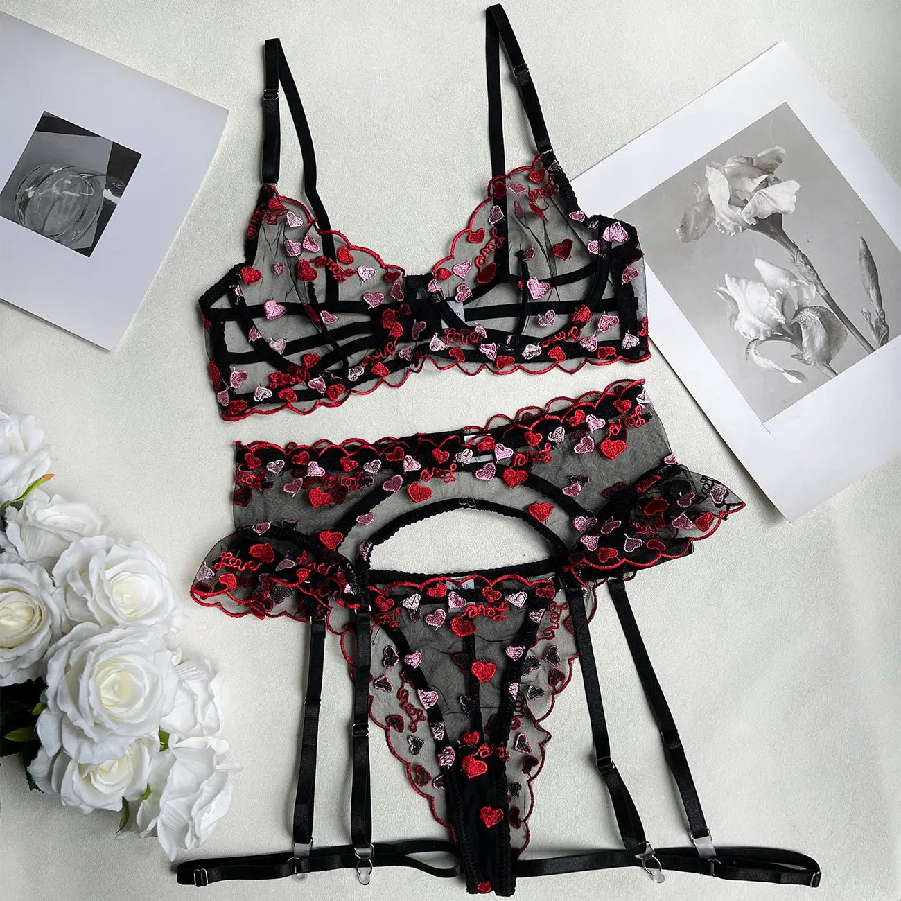 Black lingerie set with red heart details on a light surface with flowers and photos.