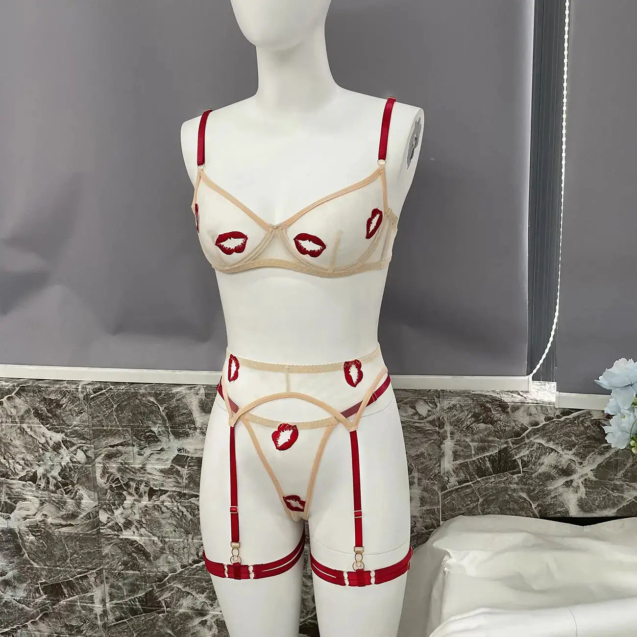 A beige sheer lingerie set featuring a demi-cup bra, garter belt, and thong, all adorned with playful red lipstick kiss embroidery and accented with red straps.
