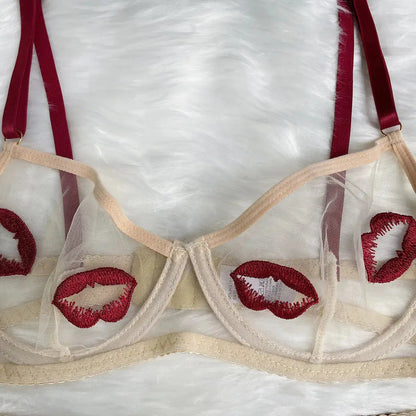 A sheer beige mesh bra featuring playful red embroidered lips and contrasting burgundy adjustable straps, laid on a soft white furry surface.