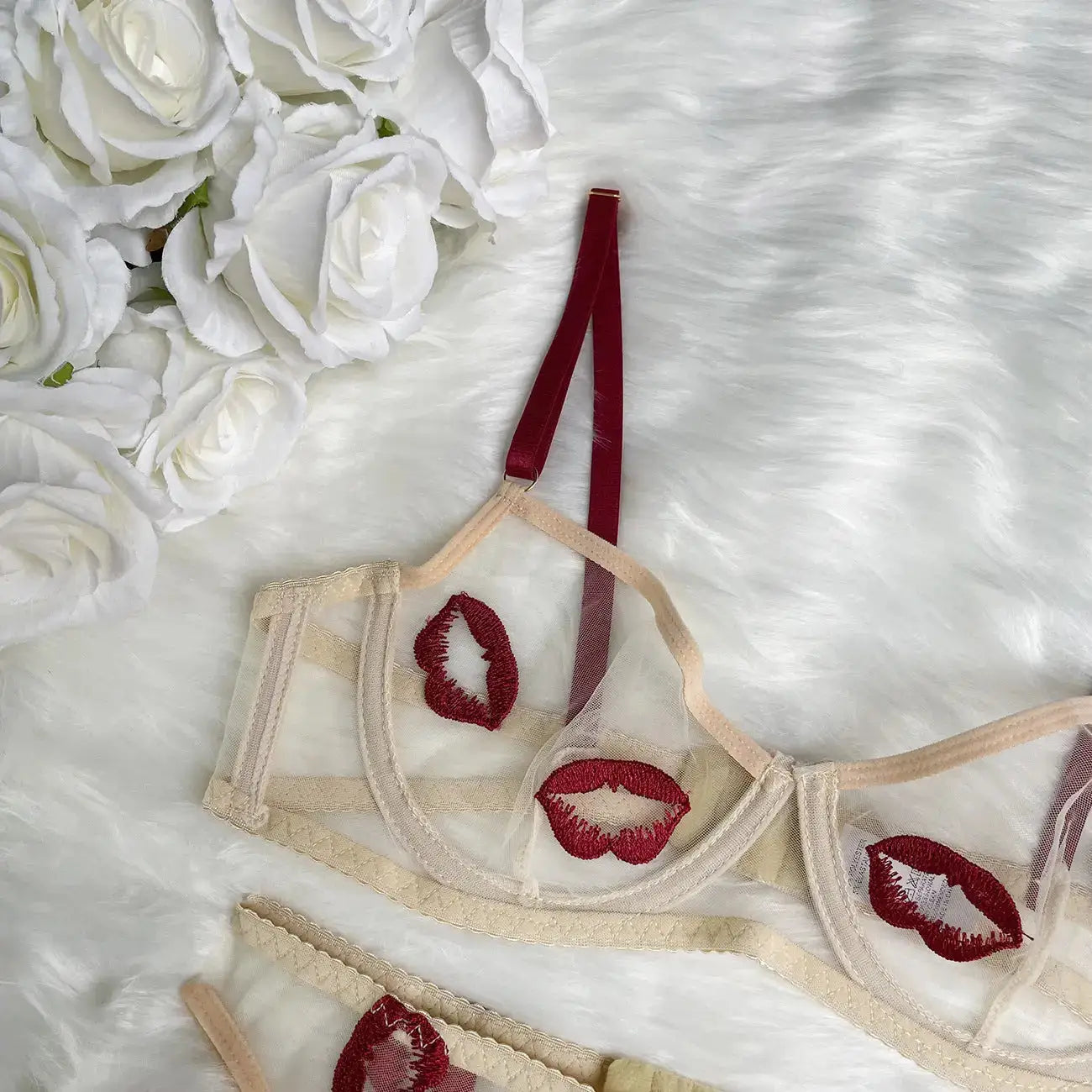 A delicate beige sheer lingerie set featuring red embroidered lips, red straps, and lace detailing, displayed on a fluffy white surface with white roses.