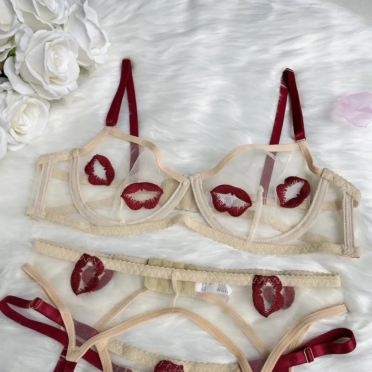 A sheer beige lingerie set featuring a bra and matching panty, adorned with red embroidered lips and burgundy straps, laid on a white furry background with white roses.