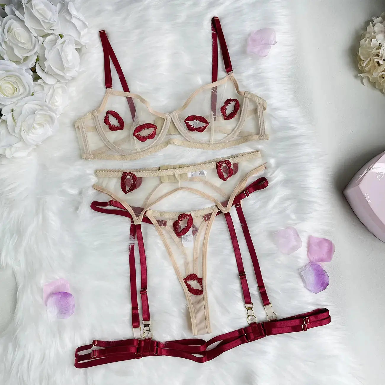This playful lingerie set includes a sheer beige mesh bra, thong, and garter belt. It features intricate red lip embroidery and adjustable deep red satin straps, laid on a white fur background.