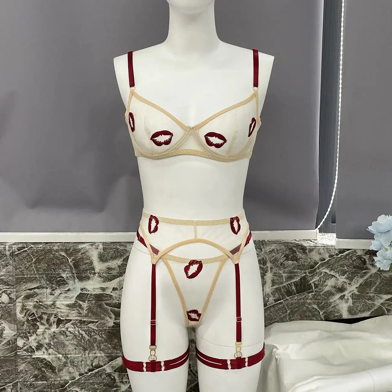 A cream sheer lingerie set featuring red lip embroidery, bra, thong, and garter belt with dark red straps, displayed on a mannequin.
