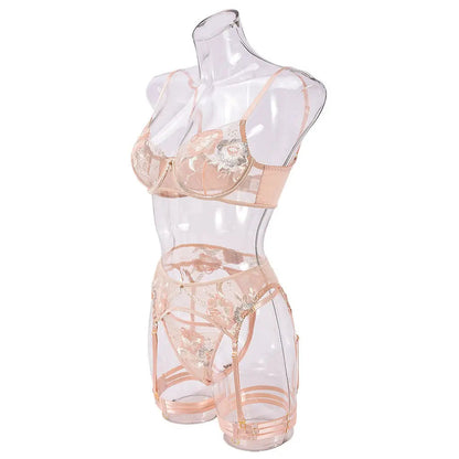 A delicate peach-colored lingerie set, featuring an embroidered bra, thong, and garter belt with intricate floral patterns, displayed on a transparent mannequin.