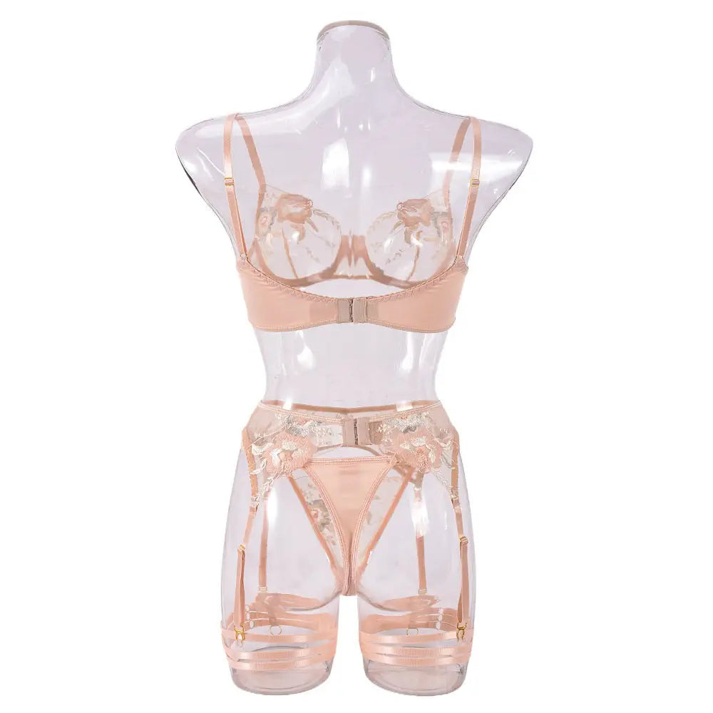 A delicate beige lingerie set featuring a sheer, embroidered open-cup bra, matching thong, and an intricate garter belt with floral lace details, displayed on a clear mannequin.