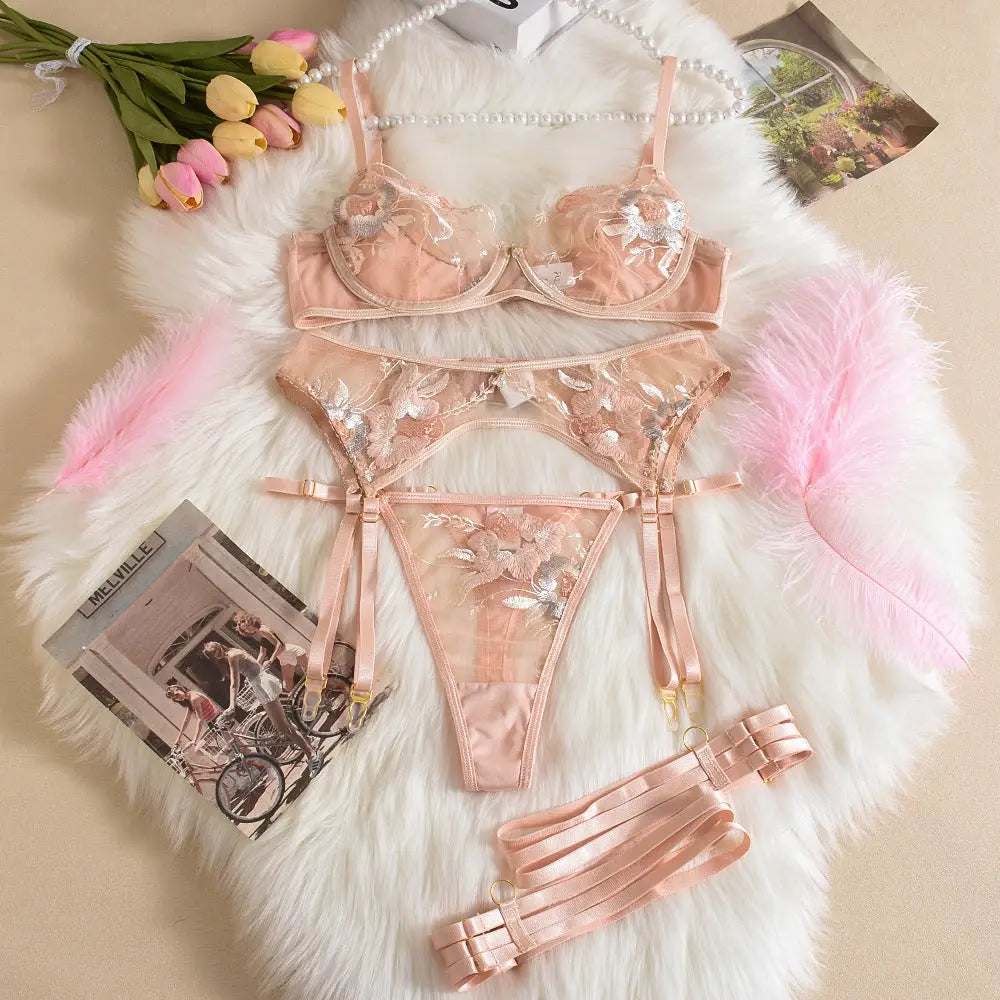 A delicate, peach-colored lingerie set featuring a bra, garter belt, and thong. It showcases intricate floral embroidery on sheer fabric, accented with metallic silver details, laid on a white furry background.