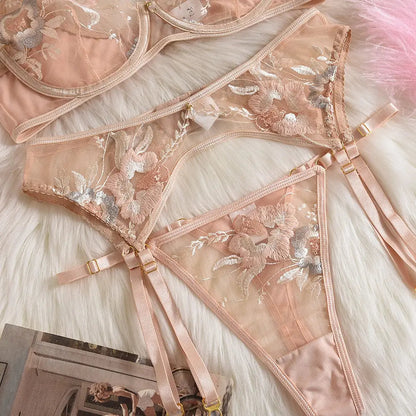 A delicate peach lingerie set featuring a bra, garter belt, and thong. It showcases exquisite floral embroidery in pink and silver on sheer mesh, laid on a white furry surface.