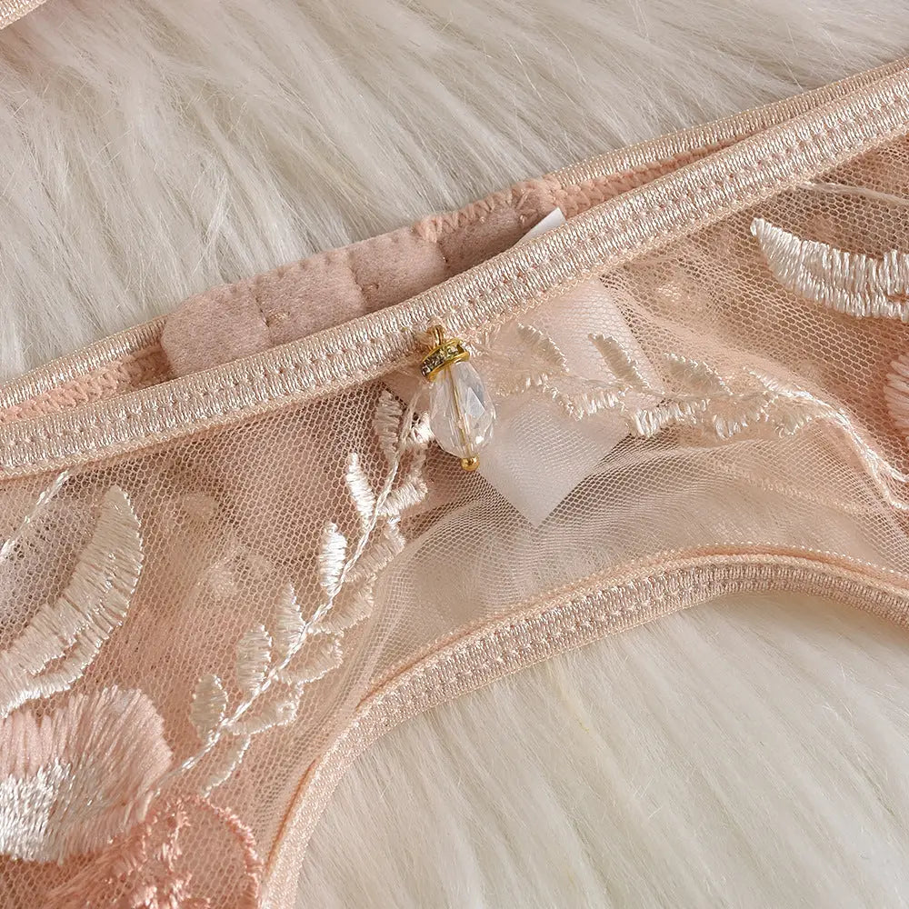 A close-up of delicate pink lace lingerie, featuring intricate white floral embroidery and a small decorative faceted charm.