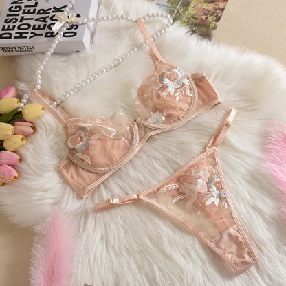 A delicate peach-colored lingerie set, featuring a sheer bra and thong with intricate floral embroidery, displayed on a plush white furry surface.