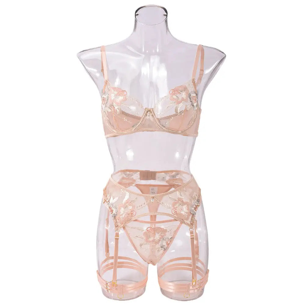 A delicate peach lingerie set features a sheer bra, thong, and garter belt, all adorned with intricate floral embroidery. It is displayed on a transparent mannequin.