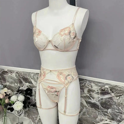 An elegant beige lingerie set, featuring a bra, garter belt, and panties, adorned with intricate floral embroidery in pink and peach hues. Displayed on a mannequin, showcasing delicate design.