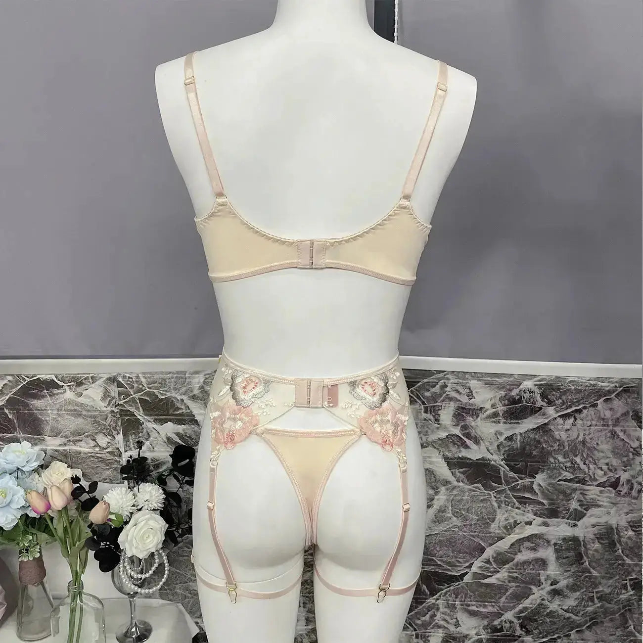 A beige lingerie set featuring a bra, thong, and garter belt with intricate pink floral embroidery and adjustable straps, displayed from the back on a mannequin.