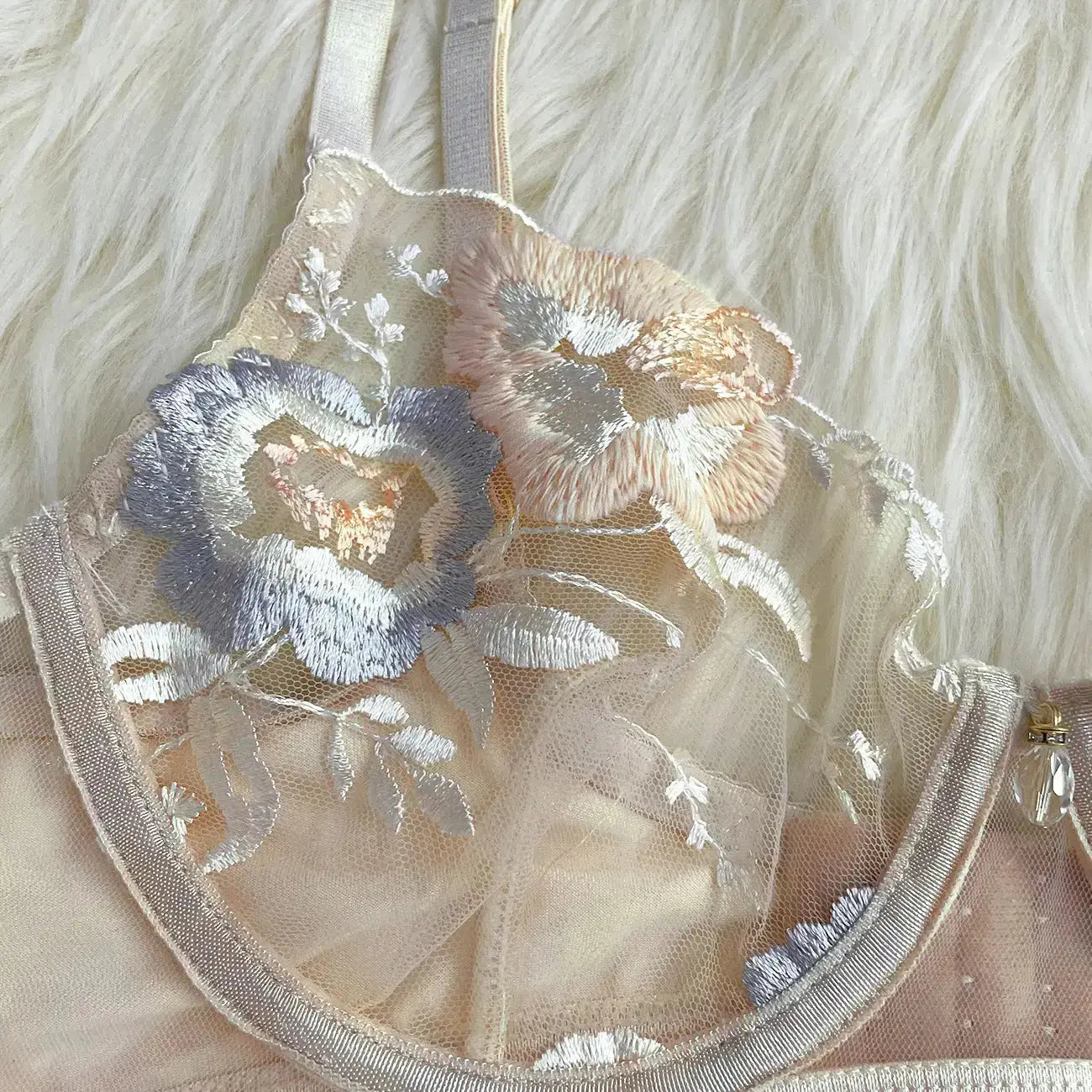 A delicate peach-colored bra, featuring sheer floral lace with intricate pastel blue, pink, and white embroidery. It has thin straps and a small decorative charm.