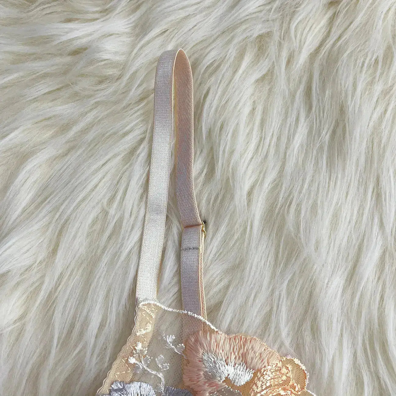 A delicate lingerie piece featuring intricate floral embroidery in pastel tones on sheer fabric, with a light pink adjustable strap, presented on a soft white fur-like background.