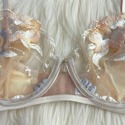 A delicate peach-toned mesh bra with intricate floral embroidery in peach, white, and light blue. It features a decorative crystal charm at the center gore, lying on a soft white background.