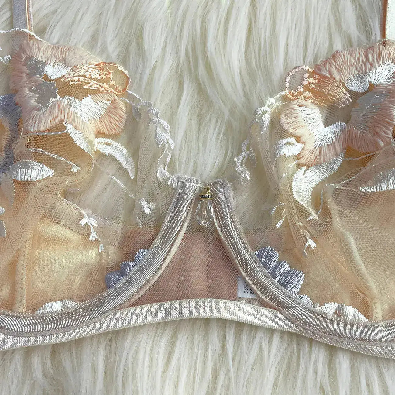 A delicate peach-toned mesh bra with intricate floral embroidery in peach, white, and light blue. It features a decorative crystal charm at the center gore, lying on a soft white background.