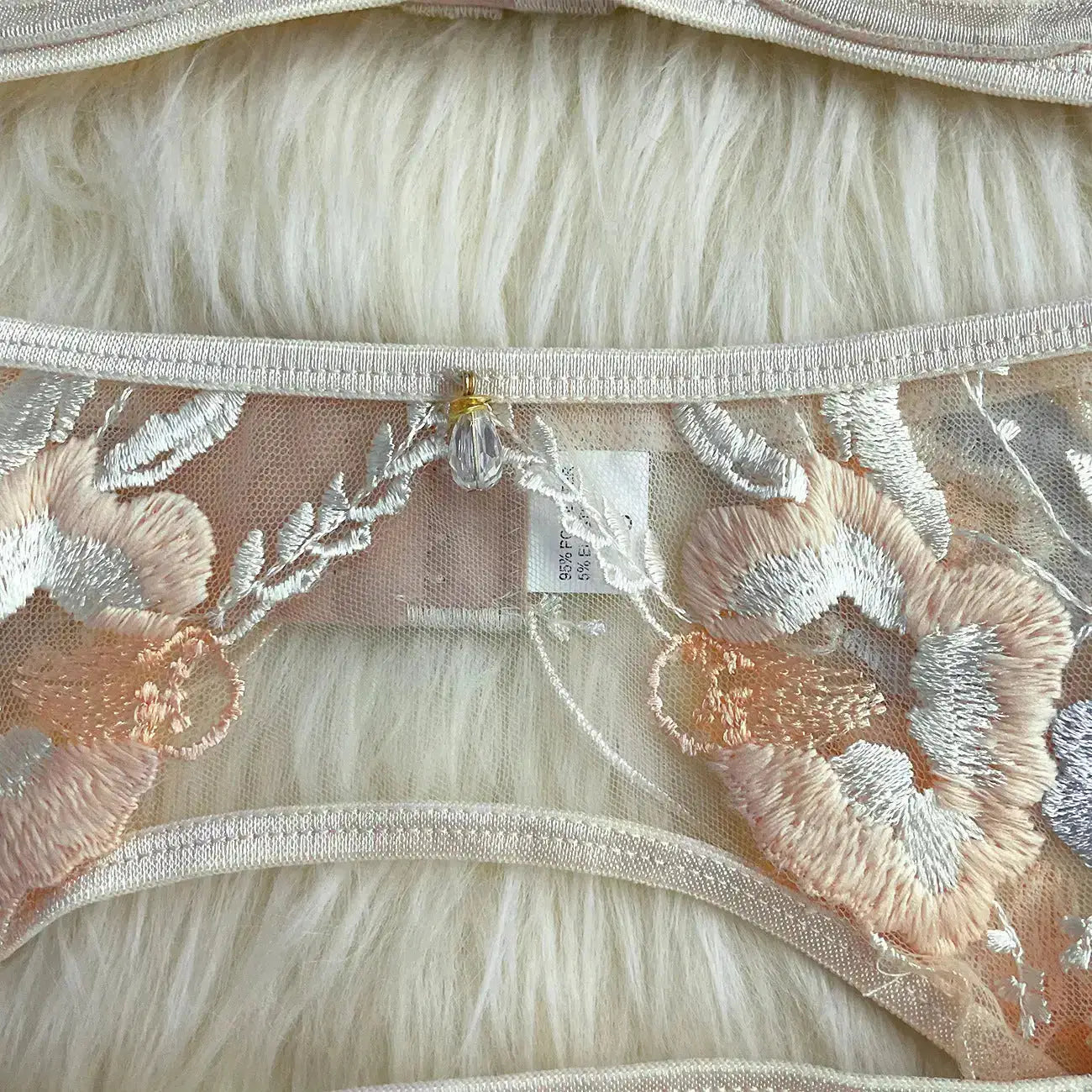Close-up of delicate peach and white floral embroidered lace lingerie on sheer fabric. It features a decorative bead and ribbed elastic trim, hinting at luxurious detail.