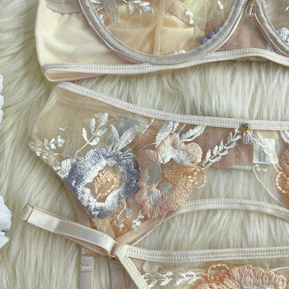 A delicate lingerie set featuring a bra and garter belt, crafted from sheer nude mesh with intricate floral embroidery in pastel peach, light blue, and silver, laid on a white fur background.