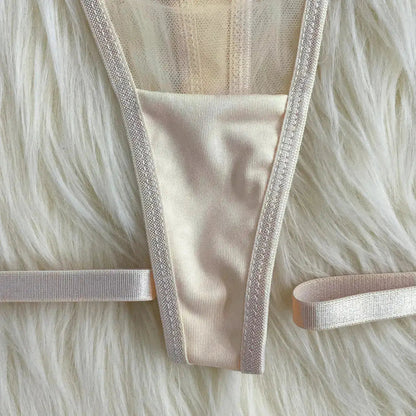 A light-colored, possibly champagne or ivory, thong-style underwear with delicate shimmering trim, laid flat on a soft, fluffy white surface. It features thin elastic side straps.