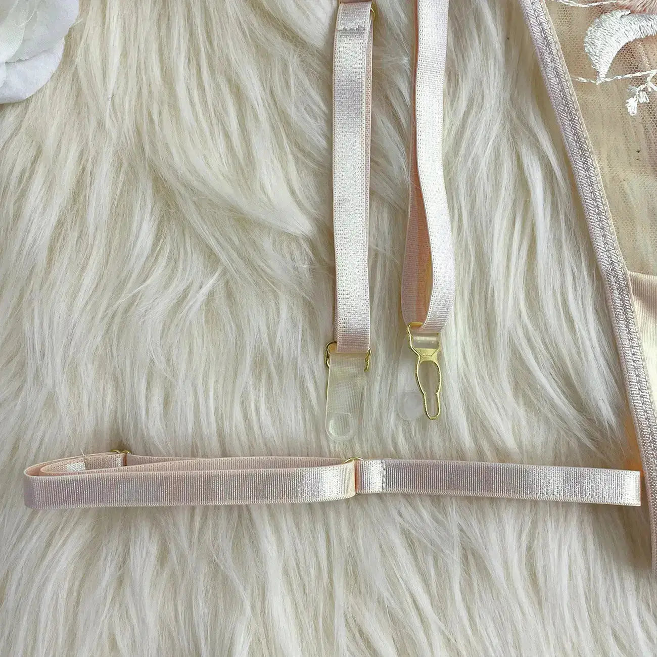 Close-up of pale pink lingerie details, featuring delicate embroidered lace, adjustable elastic straps with golden hardware, and clear garter clips, all set against a soft, furry background.