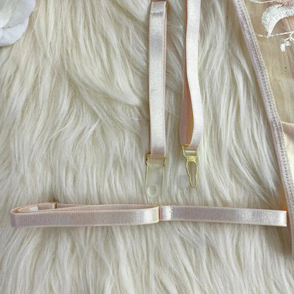 Close-up of pale pink lingerie details, featuring delicate embroidered lace, adjustable elastic straps with golden hardware, and clear garter clips, all set against a soft, furry background.
