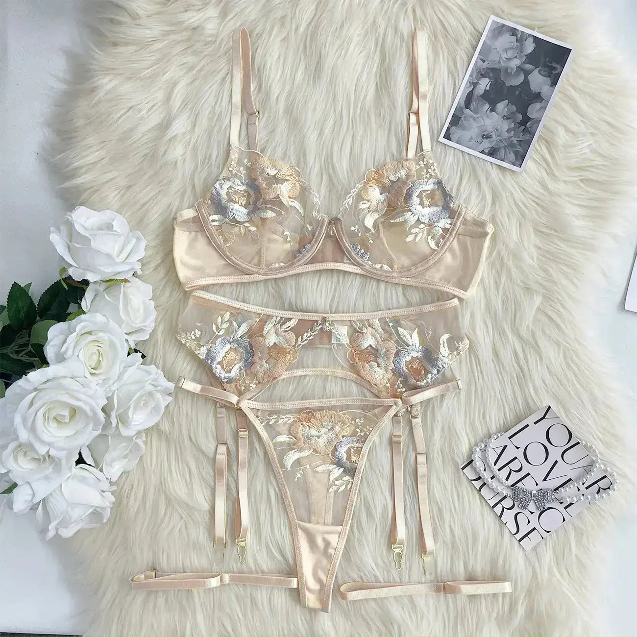 A delicate three-piece peach lingerie set featuring a sheer bra, thong, and garter belt, all adorned with intricate floral embroidery in blue, gold, and white hues.