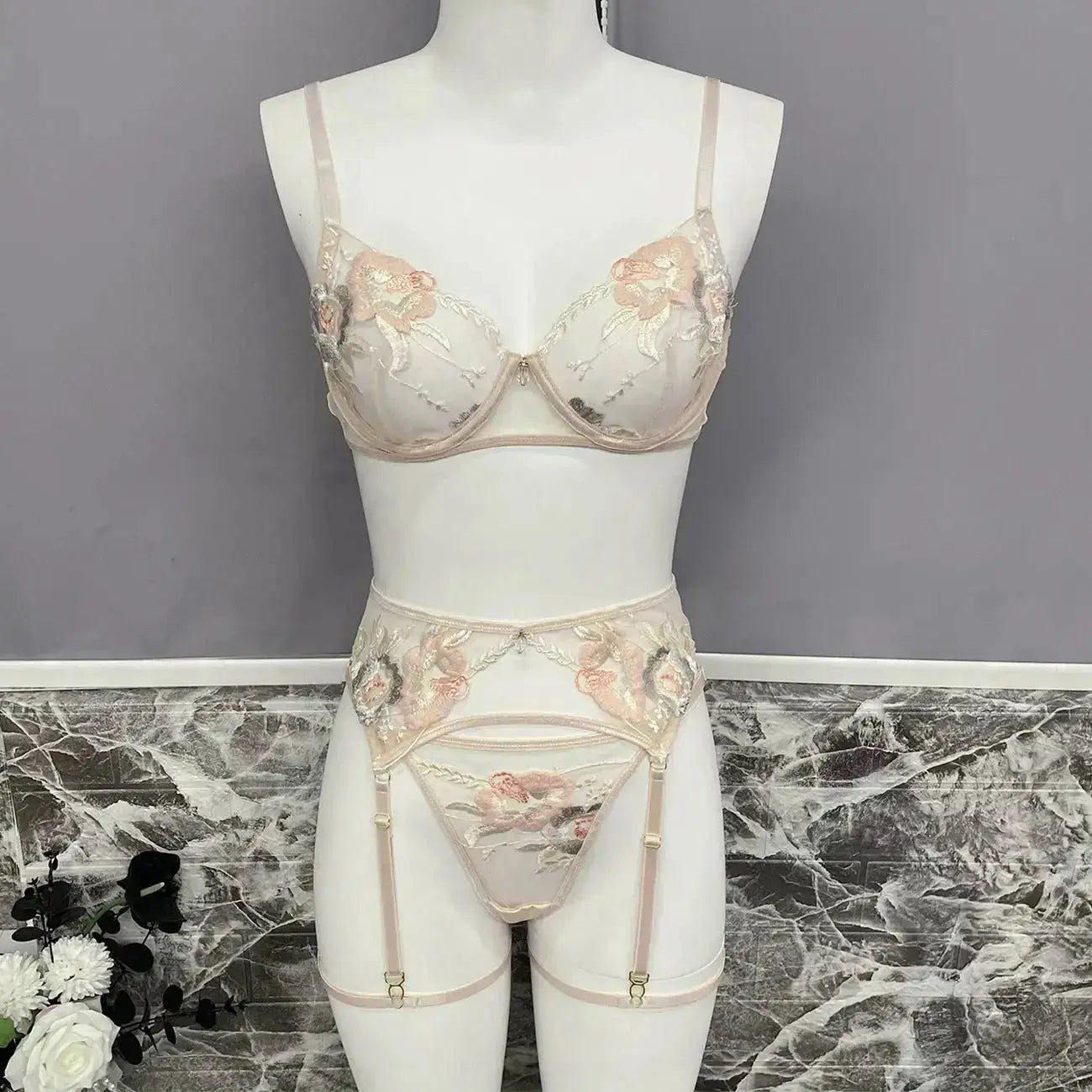 Elegant light-colored lingerie set featuring a bra, thong, and garter belt. It boasts delicate peach and sage floral embroidery on sheer fabric, displayed on a mannequin.