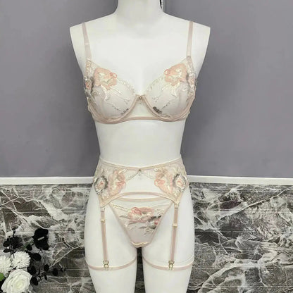 A delicate light pink lingerie set featuring a bra, thong, and garter belt, adorned with intricate floral embroidery in shades of peach, cream, and grey, displayed on a mannequin.